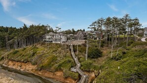 Uma área residencial costeira com casas, uma escadaria de madeira que desce até um rio, e floresta densa.