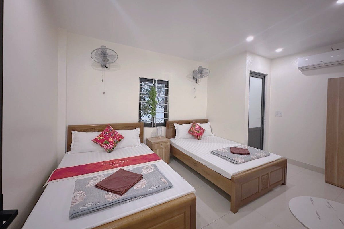 A hotel room with two single beds, each with a wooden headboard, white bedding, and red decorative pillows. There are two bedside tables with lamps, a window with a view of trees, and an air conditioning unit on the wall.