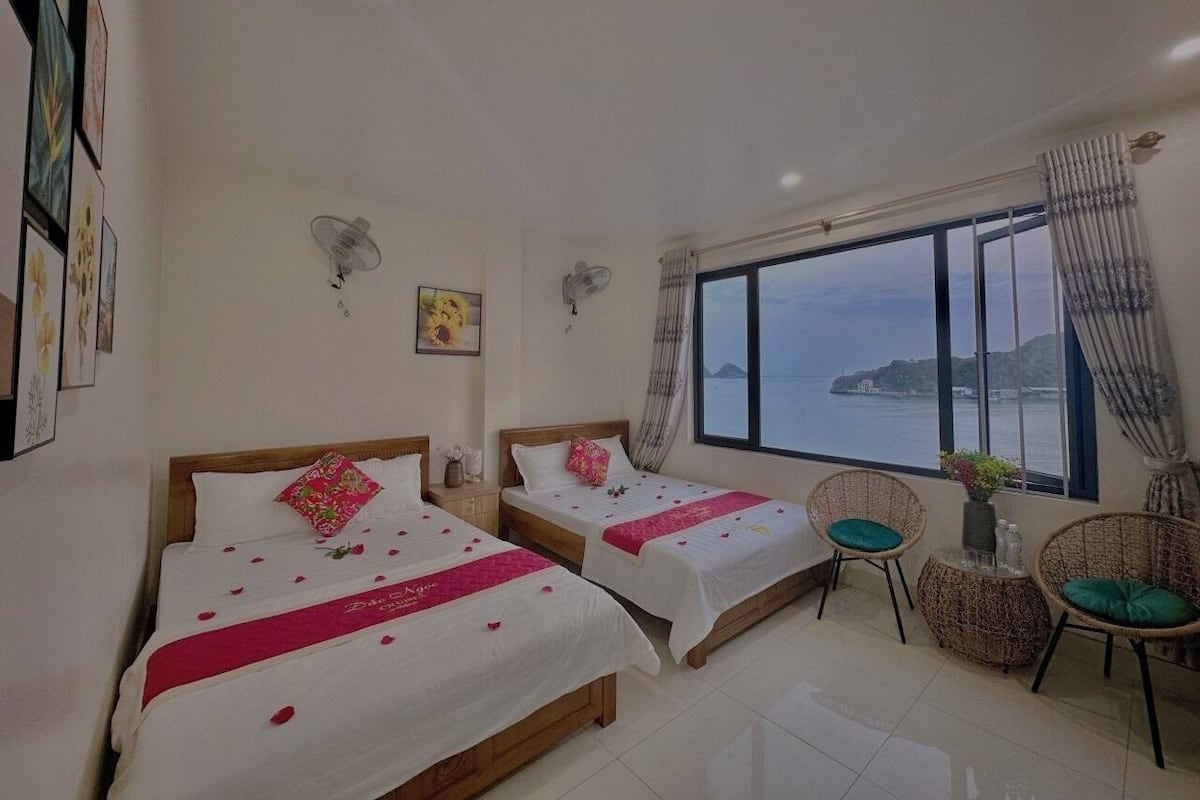 A hotel room with two beds, a large window with a view of the sea, a small table, and wicker chairs.