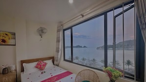 A bedroom with a bed, a chair, a nightstand, a window with a view of the sea, and a wall-mounted fan. - Gem Hostel (Hai Phong)