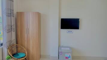 Ein Zimmer mit Holzschrank, Korbsessel, kleinem Tisch mit Pflanze, Kühlschrank, an der Wand montiertem Fernseher und Schild zur Kennzeichnung der WLAN-Verfügbarkeit.