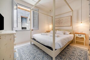 A four-poster bed with white linens, a bedside table with a lamp, a painting on the wall, and a patterned floor.