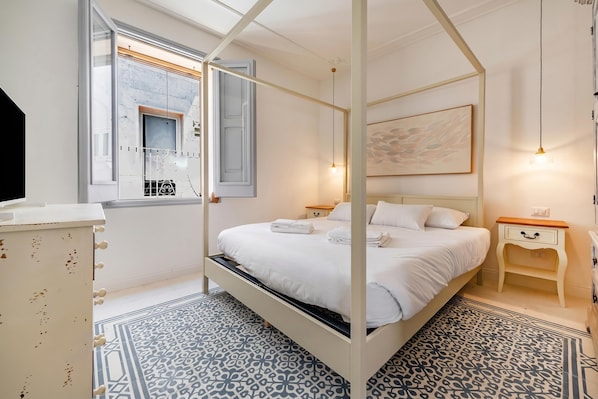 A four-poster bed with white linens, a bedside table with a lamp, a painting on the wall, and a patterned floor.