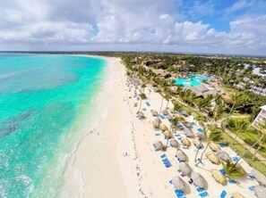 A beach resort with a clear turquoise sea, white sand, and a pool area with umbrellas and lounge chairs.