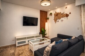 A modern living room with a flat-screen TV, a white entertainment unit, a dark sofa, a glass coffee table, a wooden accent wall, and a decorative map.