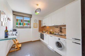 A compact kitchen with a washing machine, a wooden countertop, and a window with colorful blinds.