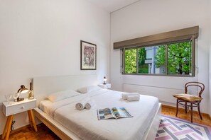 A modern bedroom with a large bed, a bedside table with a lamp, a chair, and a window with a view of greenery.