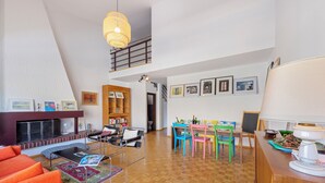 Un amplio salón con techo alto, chimenea, estantería y zona de comedor con sillas de colores.