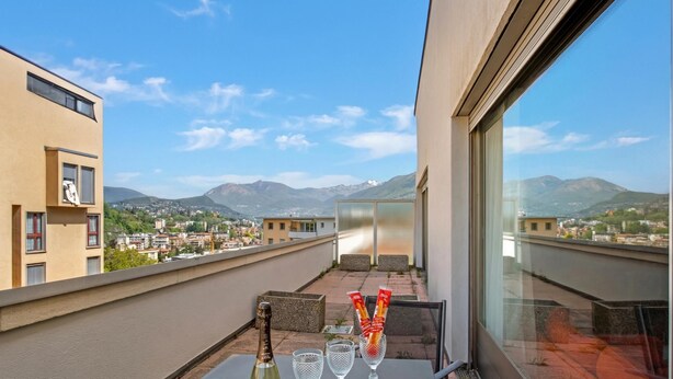 Un balcón con una mesa, una botella y vasos, con vistas a una ciudad y montañas.