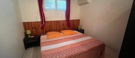 A bedroom with a bed, two bedside tables, a window with curtains, and an air conditioning unit.