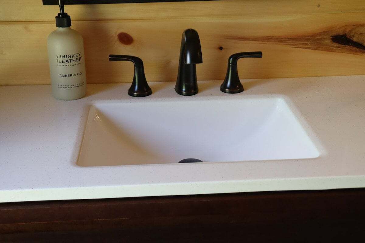 Ein Badezimmerwaschbecken mit weißer Ablage, einer Whiskeyflasche, Lederseifen und drei schwarzen Wasserhähnen.
