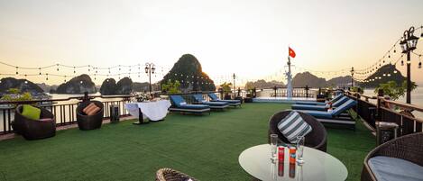 A rooftop terrace with wicker furniture, a table with drinks, and a view of the water and mountains.