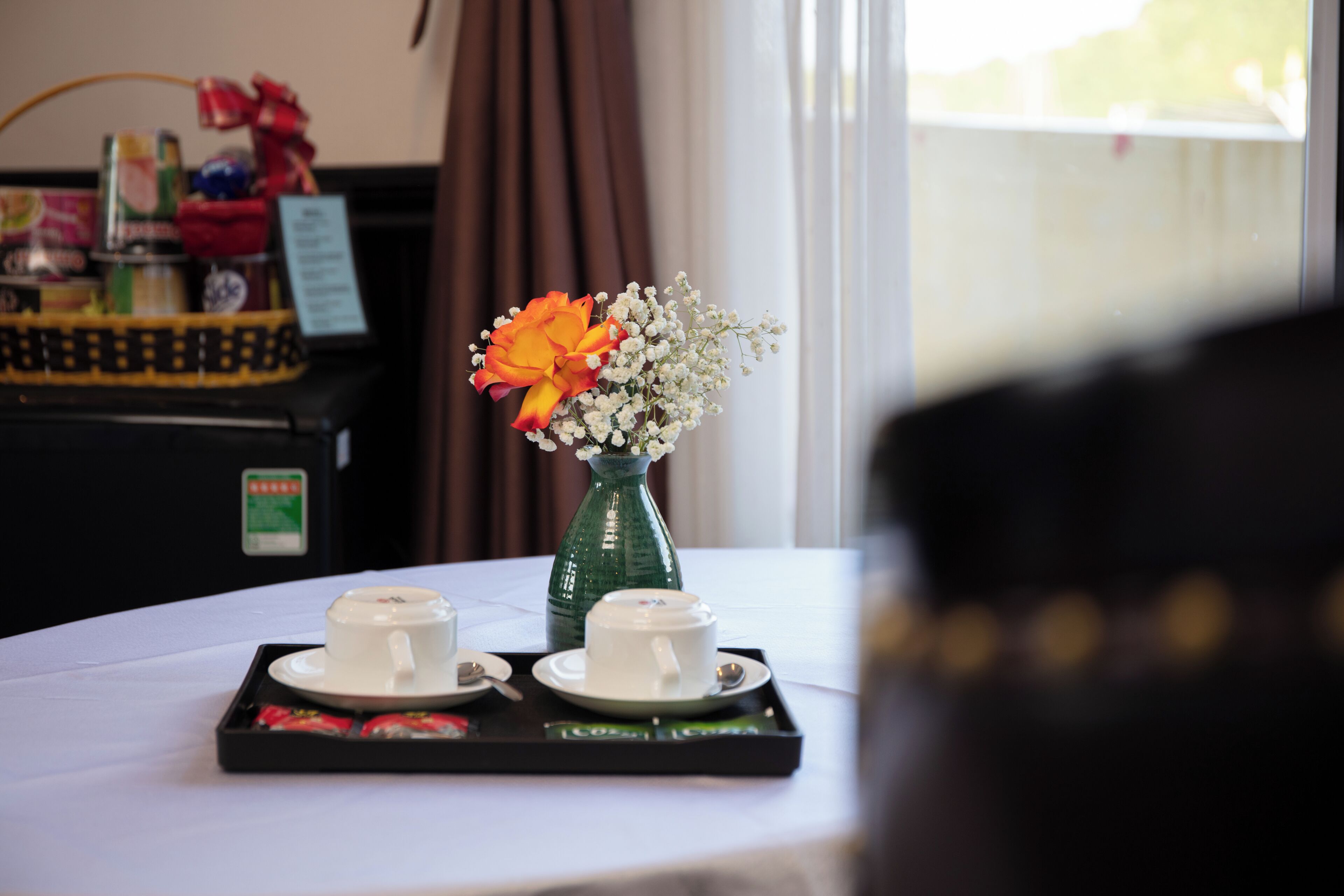 Una mesa con dos tazas blancas y una bandeja de pasteles, un jarrón verde con flores, y un tablero de menú al fondo.