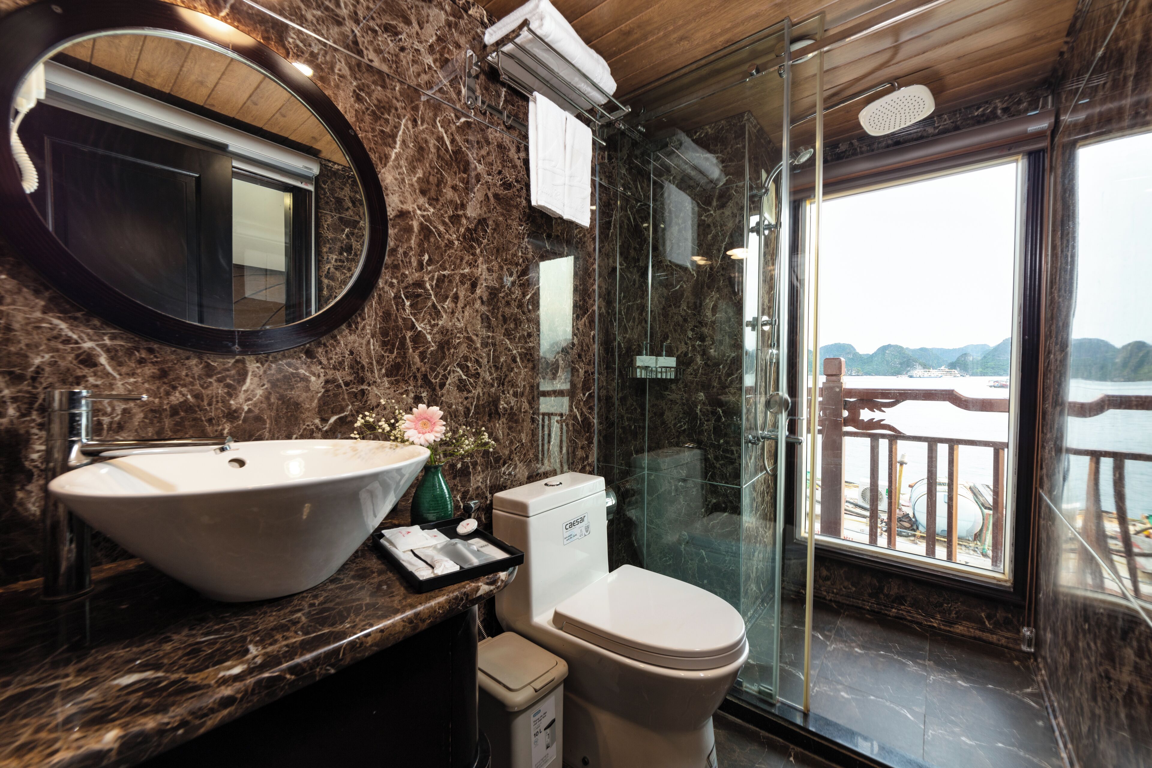 A bathroom with a marble sink, toilet, and a large mirror. There is a window with a view of the sea and a balcony.