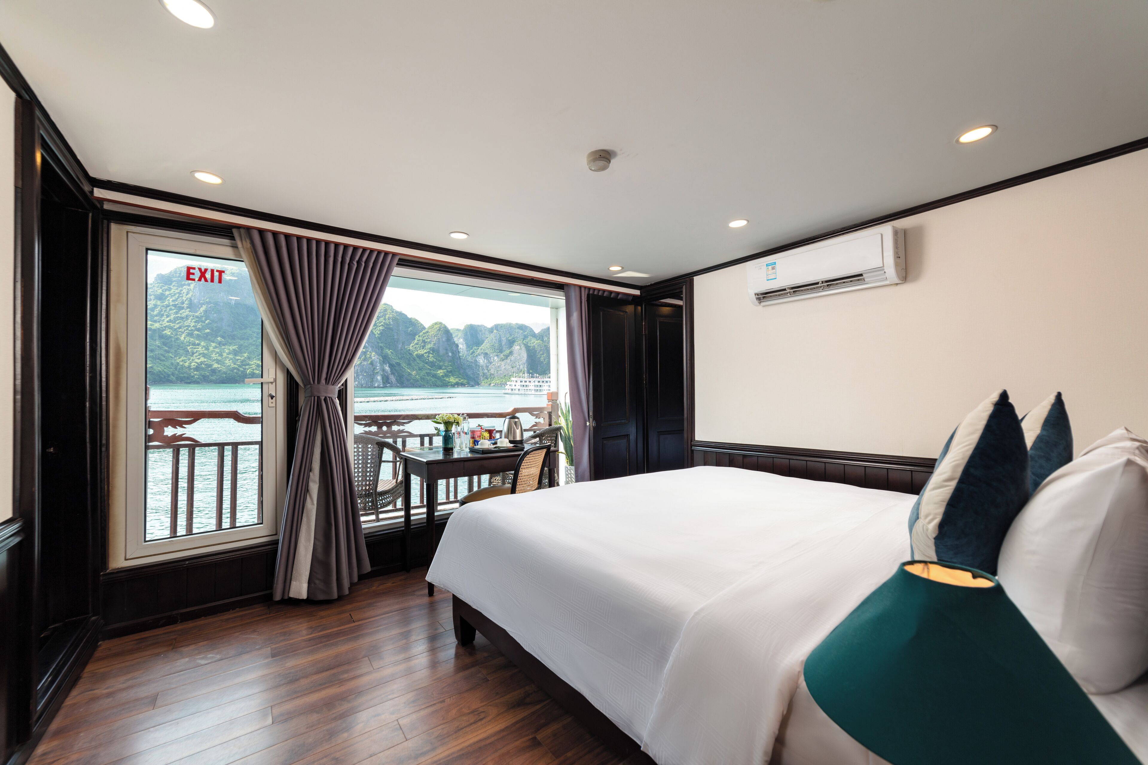 A hotel room with a large bed, a table with chairs, a view of the sea, and an exit sign.