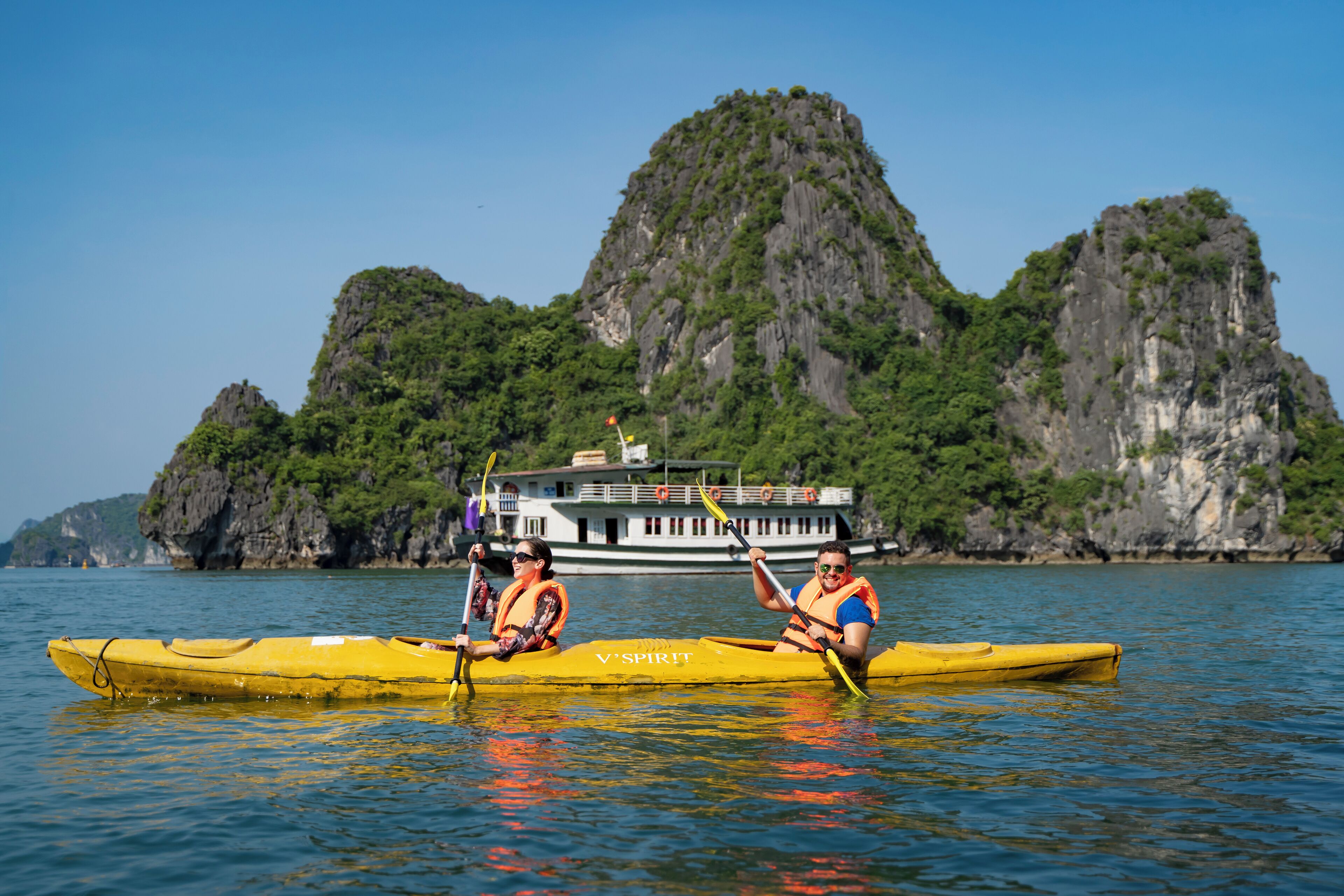Two people in a yellow kayak on calm water with a boat and rocky cliffs in the background.