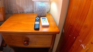 Een houten nachtkastje met een lamp, een afstandsbediening en een klein elektronisch apparaat.