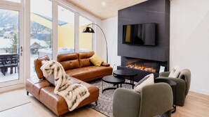 A modern living room with a fireplace, leather sofa, and a television mounted on the wall. - 4 BR with Epic Mountain View, Hot Tub, Garage EV Port, Super High Ceiling (Canmore)