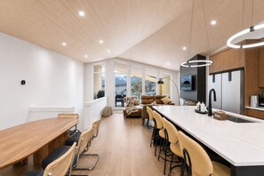 A modern kitchen with a large island, bar stools, and a dining area with a wooden table and chairs. - 4 BR with Epic Mountain View, Hot Tub, Garage EV Port, Super High Ceiling (Canmore)
