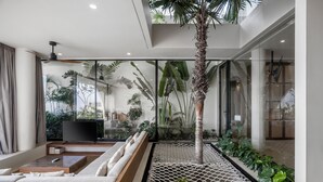A modern living room with a large TV, comfortable seating, a ceiling fan, and a view of tropical plants.