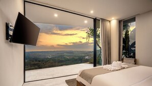 A modern bedroom with a large bed, a flat-screen TV mounted on the wall, and a view of a landscape through a sliding glass door.