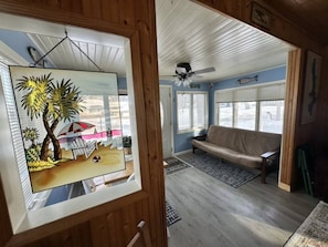 Et værelse med trælofter, en ventilator, en sofa, et vindue med udsigt og et billede af en strand.