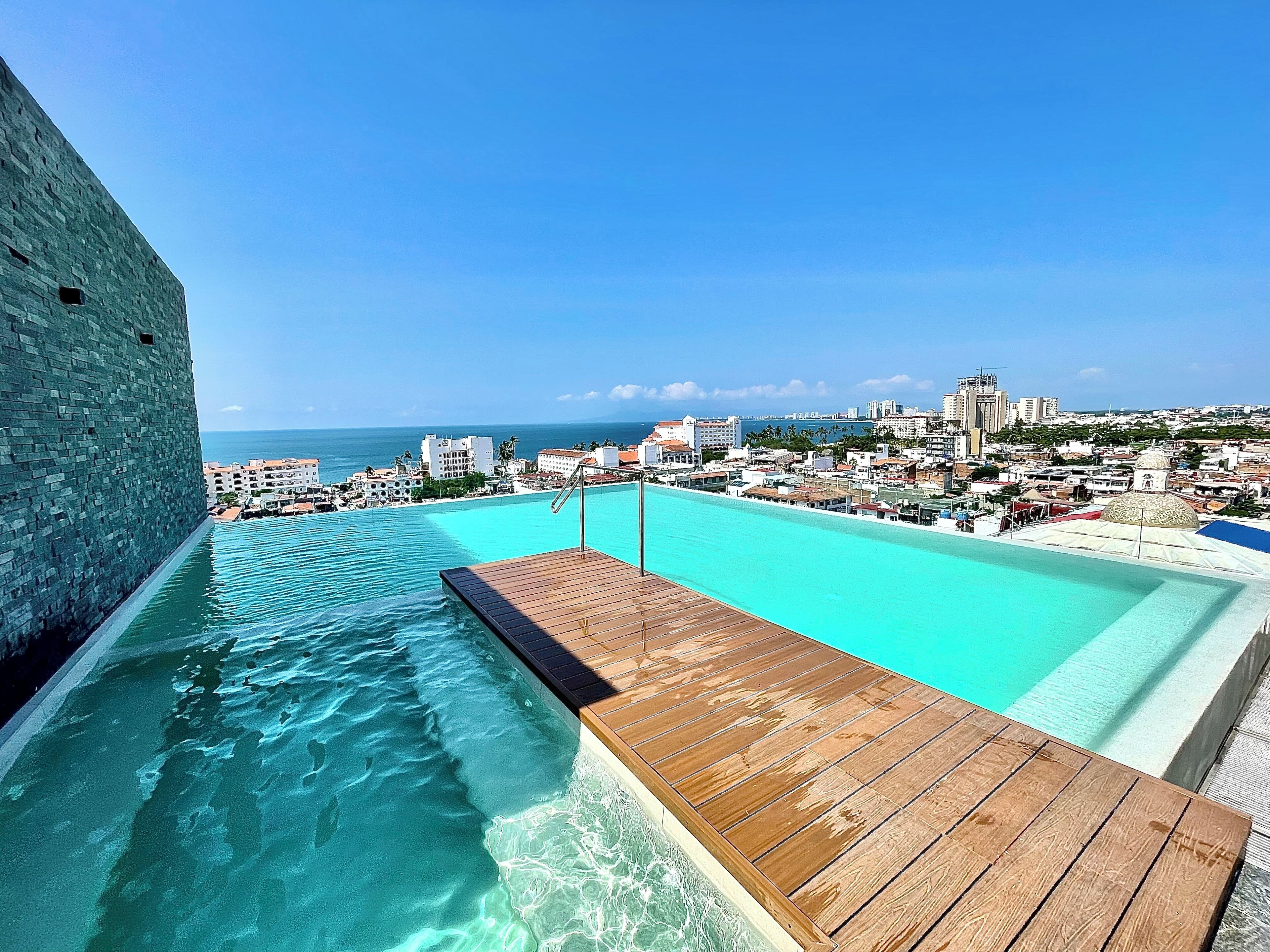 A rooftop infinity pool with a wooden deck, overlooking a coastal cityscape and the ocean.
