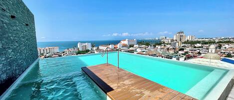 A rooftop infinity pool with a wooden deck, overlooking a coastal cityscape and the ocean.