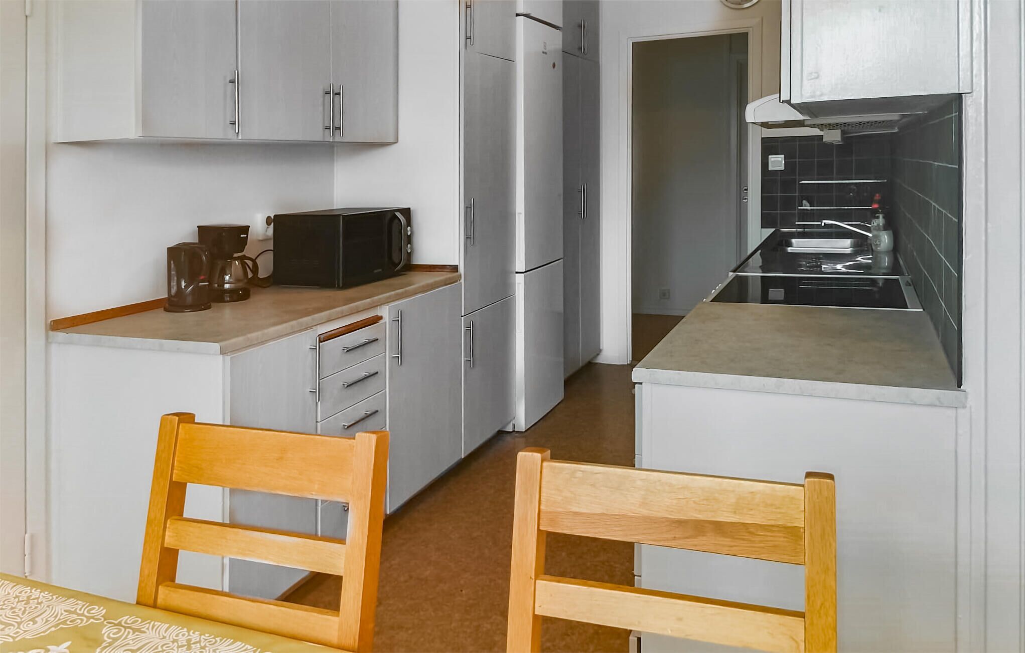 Een kleine keuken met witte kastjes, een magnetron en een koffiezetapparaat op het aanrecht. Op de voorgrond staan twee houten stoelen.
