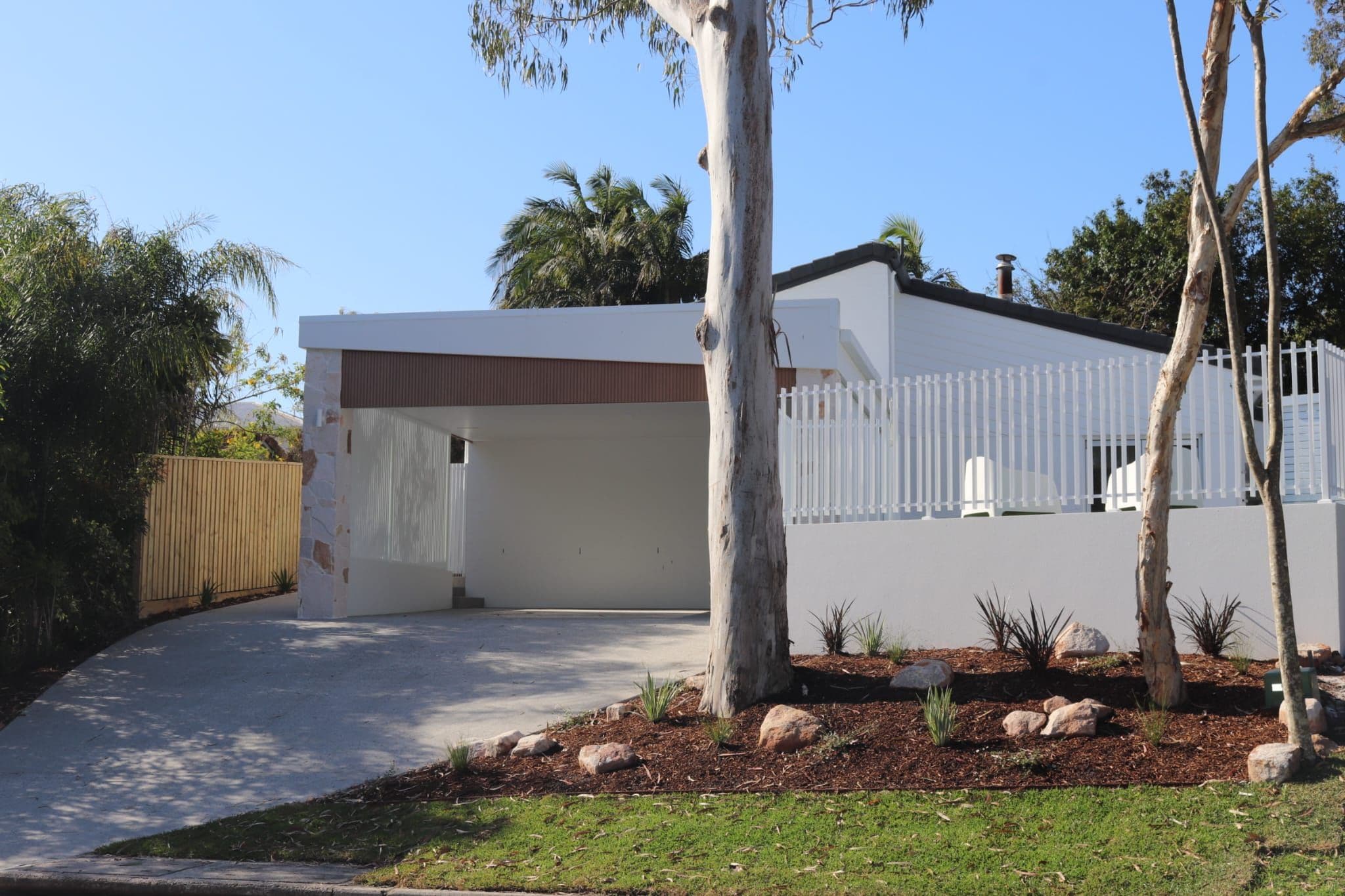 Ein modernes Haus mit weißer Fassade, einer überdachten Auffahrt und einem von Bäumen und Sträuchern umfriedeten Garten.