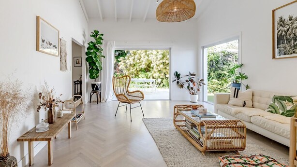 A spacious living room with a high ceiling, a large window, and a view of greenery. There are wicker furniture pieces, a sofa, and a dining table.