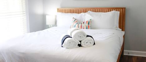Una cama bien hecha con sábanas blancas y toallas enrolladas, cabecera de madera y una sola almohada con un estampado colorido.