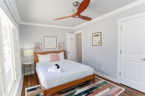 Ein Schlafzimmer mit einem Bett, einem Deckenventilator, einem Nachttisch, einer Lampe und gerahmten Kunstwerken an der Wand.