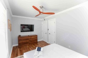 Un dormitorio con un aparador de madera, televisor montado en la pared, ventilador de techo y una cama con toallas enrolladas.