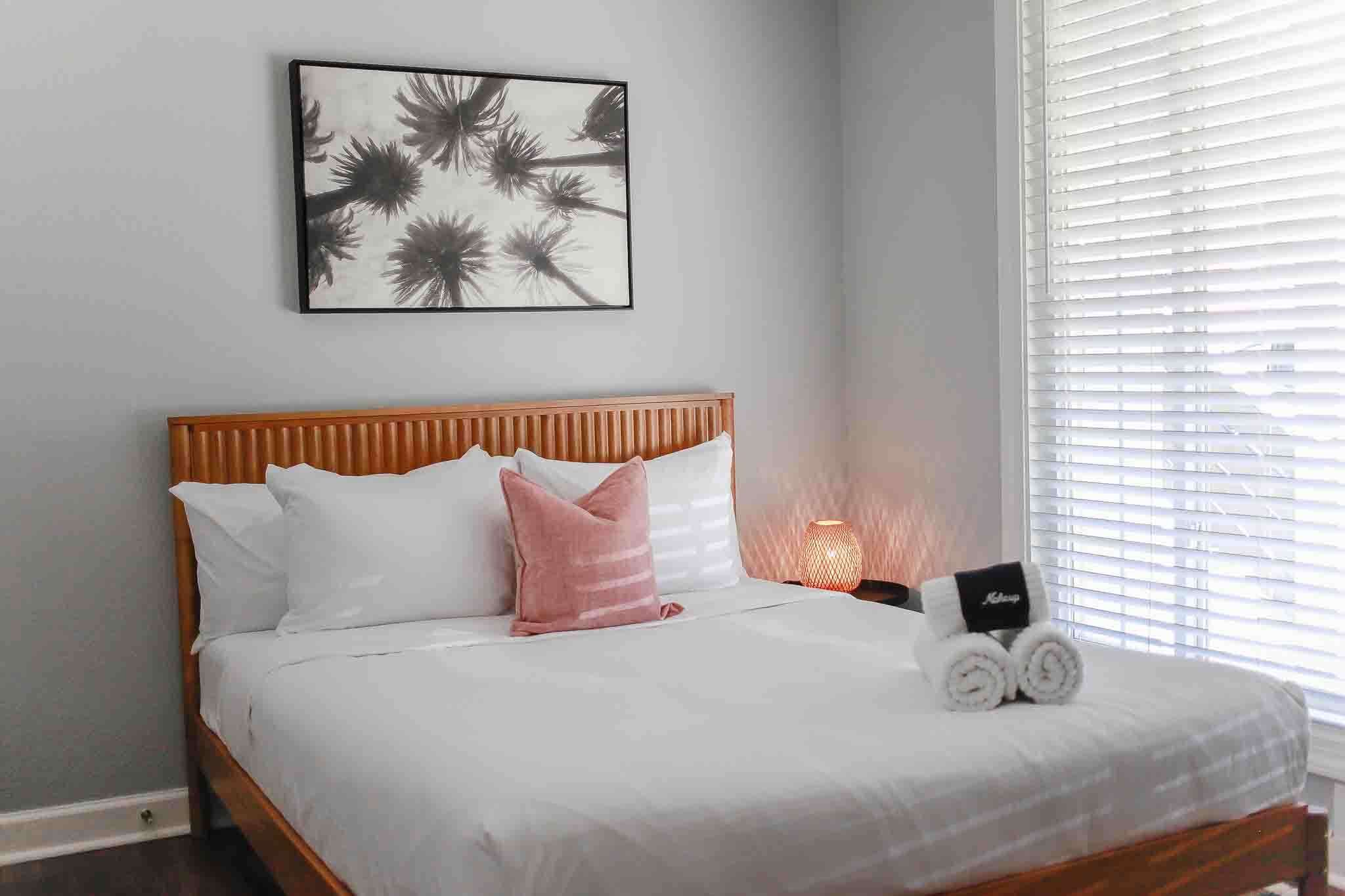 A neatly made bed with white and pink pillows, a framed artwork above it, and a bedside lamp.