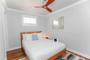 A bedroom with a wooden bed, a ceiling fan, a bedside lamp, a small table with a towel, and a window with blinds. - Sojourn Salt Suite 2 BR 2 BA at Beach Cottages (Norfolk)