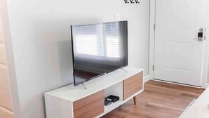 Ein modernes Wohnzimmer mit einem Flachbildfernseher auf einer weißen Ablage, Holzboden und einer weißen Tür im Hintergrund.