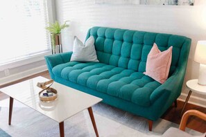A teal upholstered sofa with two pillows, a small white table with a tray, and a lamp on a side table. - Sojourn Salt Suite 2 BR 2 BA at Beach Cottages (Norfolk)