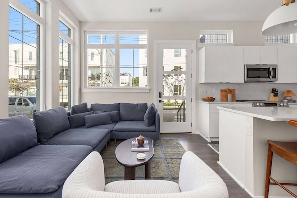 A modern living room with a grey sectional sofa, a white armchair, and a small coffee table. A kitchen with white cabinets and a microwave is visible in the background.
