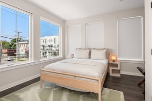 A bedroom with a large window, a bed with checkered bedding, a bedside table with a lamp, and a view of buildings outside.