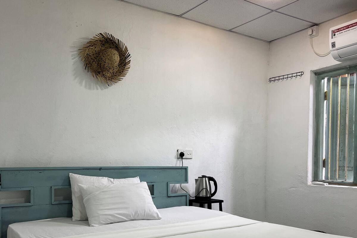 Una cama bien hecha con sábanas blancas, un cabecero verde azulado, una lámpara de pared y una ventana con barrotes.