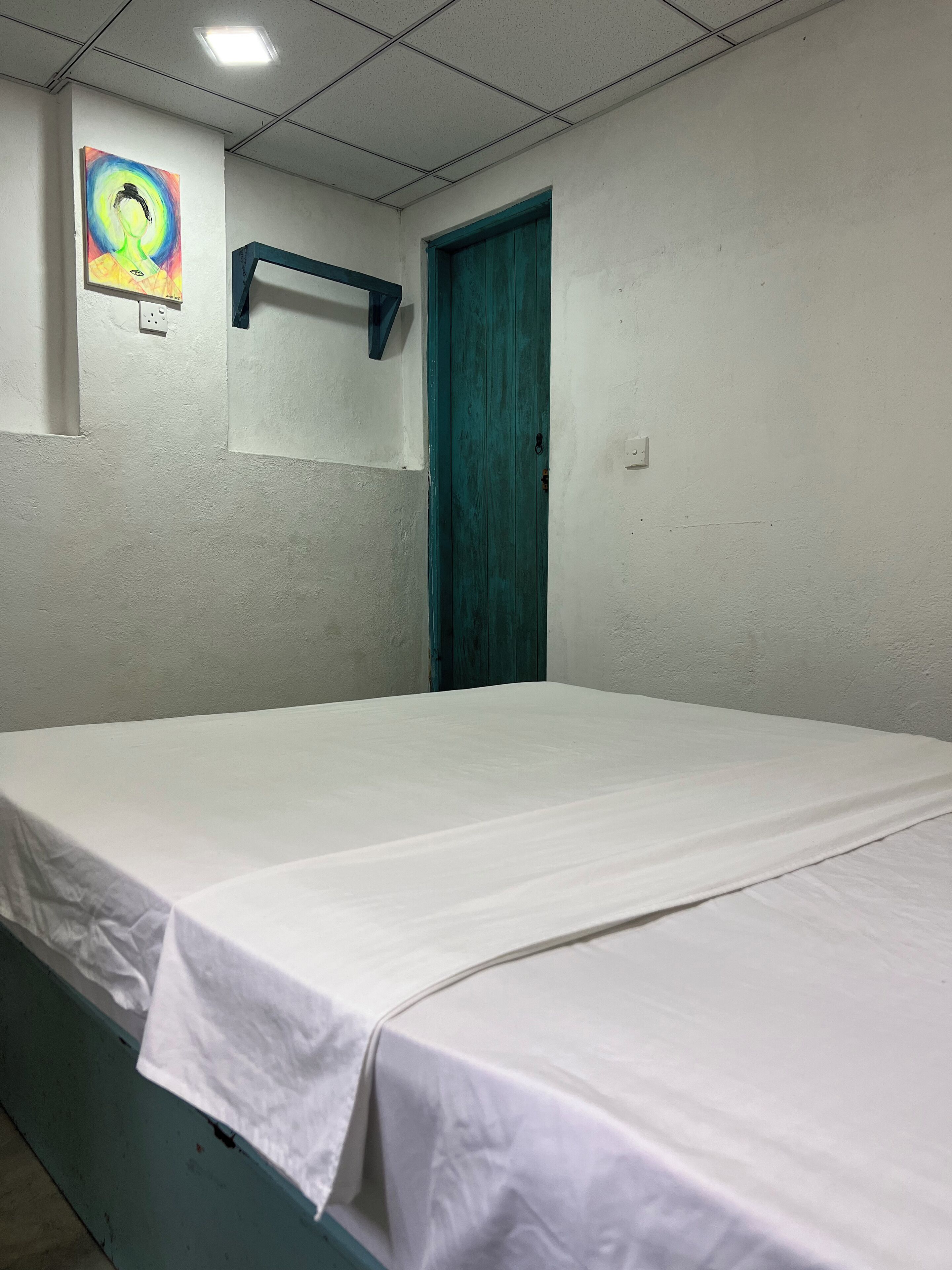 Een kamer met een enkel bed, een blauwgroene deur, een wandplank en een plafondlamp.
