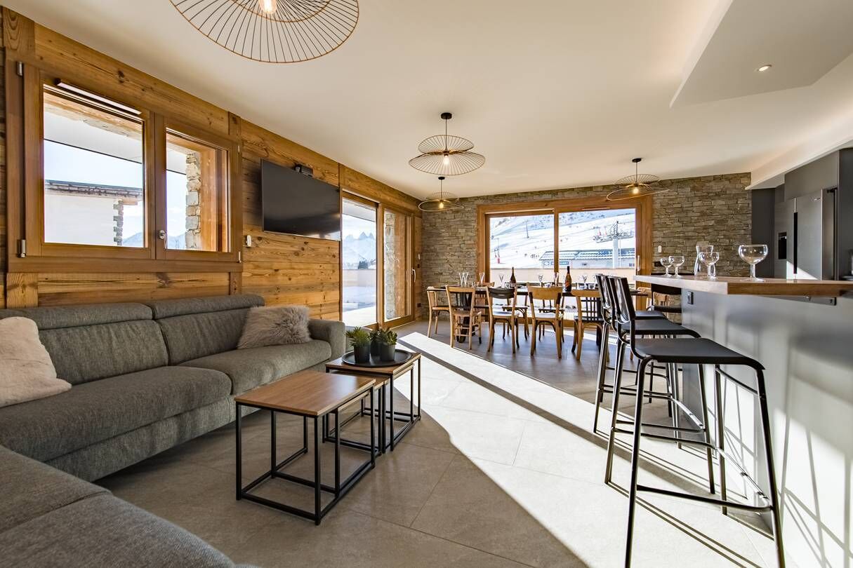 A modern interior with a wooden wall, a grey sofa, a dining area with a table and chairs, and a kitchen area with a bar.