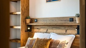 Ein Schlafzimmer mit einem hölzernen Kopfteil, einem Bett mit weißen und gemusterten Kissen, einer Wandleuchte und einem gerahmten Bild von Bergen.
