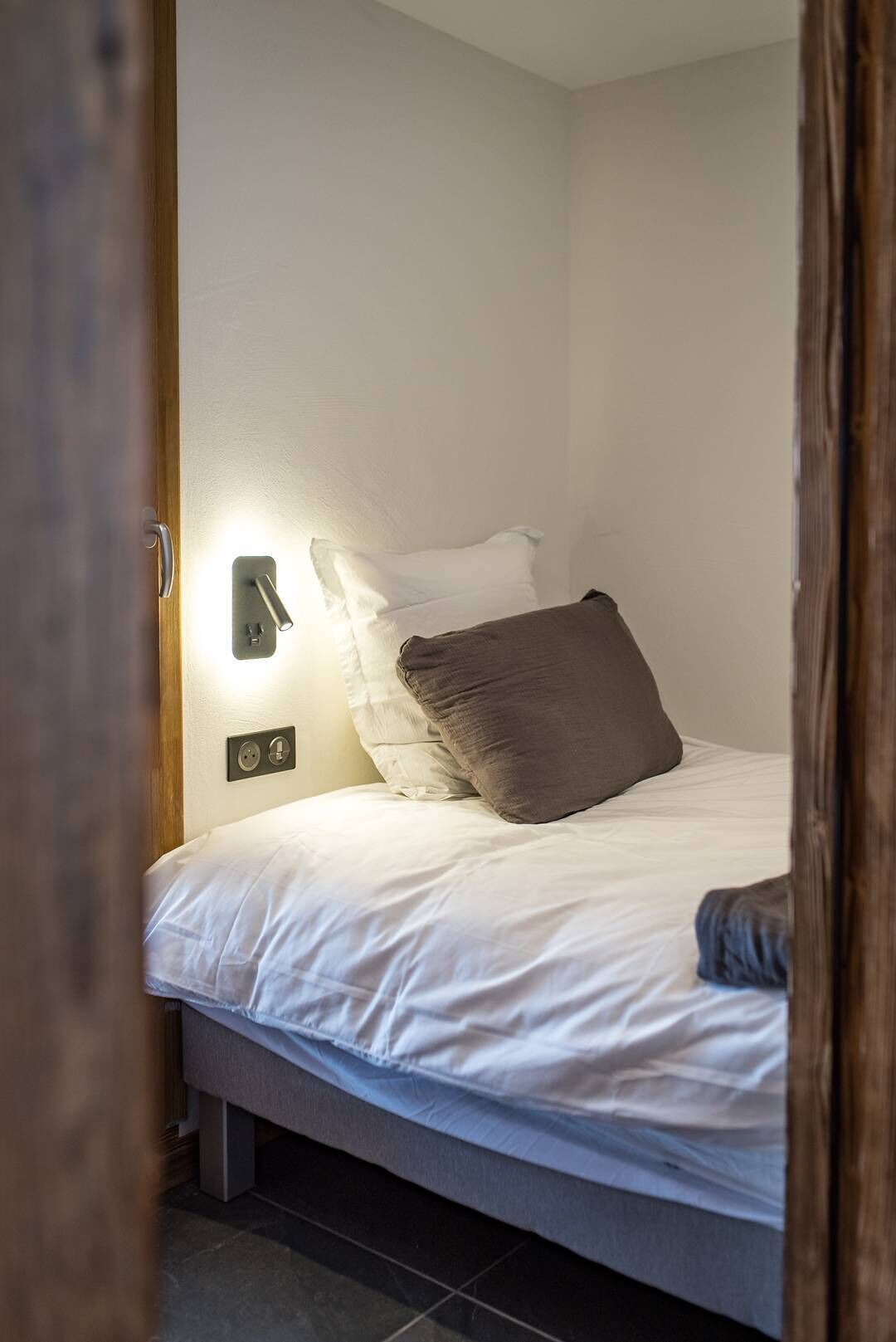 Una cama bien tendida con ropa blanca y una sola almohada oscura, un aplique en la pared y un marco de puerta visible en primer plano.