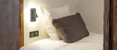 Una cama bien tendida con ropa blanca y una sola almohada oscura, un aplique en la pared y un marco de puerta visible en primer plano.