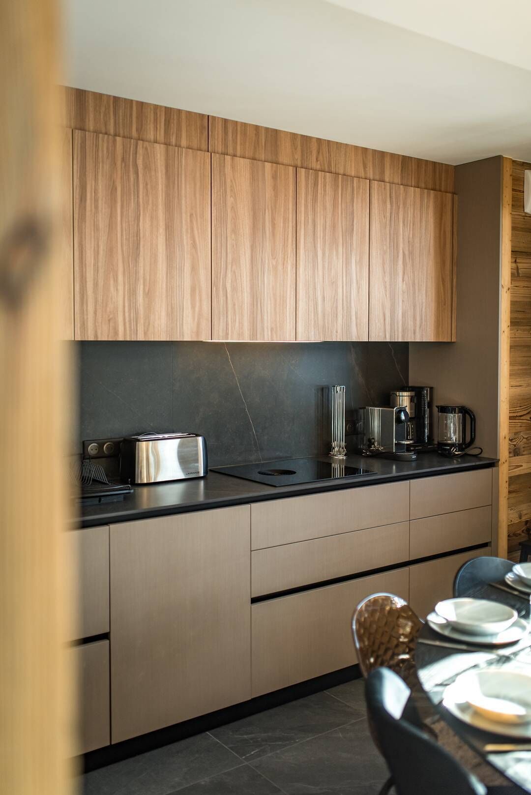Eine moderne Küche mit Holzausstattung, einer schwarzen Arbeitsfläche und einem Essbereich mit Stuhl und Geschirr.