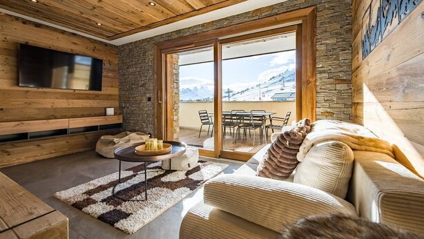 A cozy living room with a wooden ceiling, stone walls, a large TV, a sofa, a coffee table, and a view of the mountains through the glass doors. - Apartment for 6/8 people with jacuzzi in a new residence on the slopes (LA TOUSSUIRE)