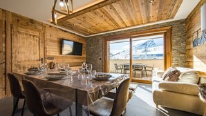 A dining area with a wooden table set for four, a stone wall, and a large window overlooking a snowy landscape. - Apartment for 6/8 people with jacuzzi in a new residence on the slopes (LA TOUSSUIRE)