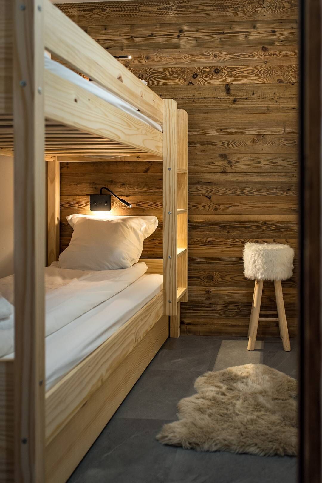 Um quarto pequeno de madeira com uma cama beliche, um banquinho e um tapete de pele.